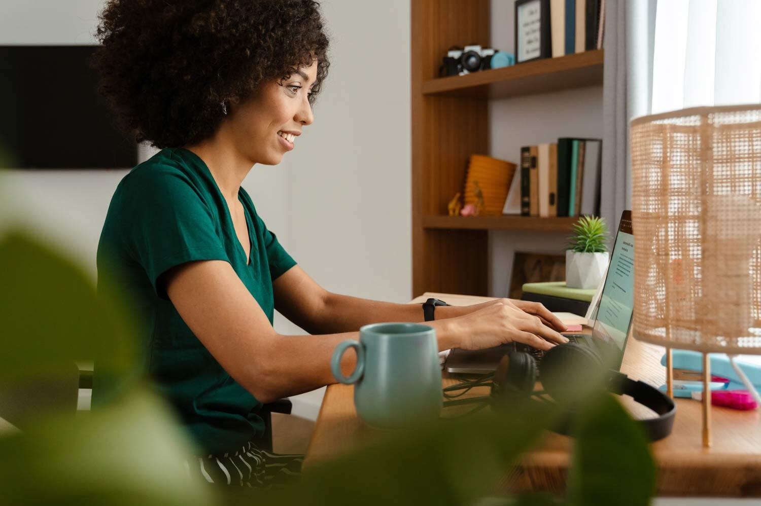 Smiling woman with curly hair typing on laptop at desk in home office, with mug, headphones, lamp and bookshelf in background