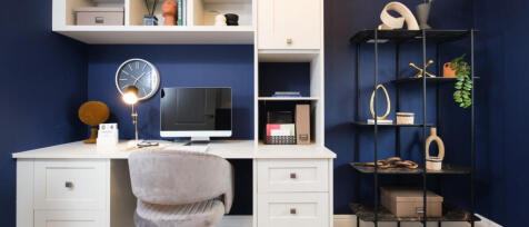 Modern home office with white desk, beige chair, navy blue wall, and black metal shelving holding de