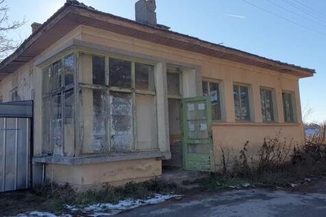 Side perspective of the old bakery showing beige walls, green door, and adjacent metal shed under a clear sky.