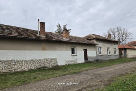Side facade of a Bulgarian house showing multiple chimneys, windows, and a dirt road alongside the building.