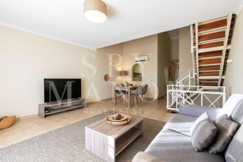 Open-plan living room in a furnished triplex apartment for sale at Gramacho Golf Resort, Carvoeiro, Algarve. Features dining area, sofa, and stairs.