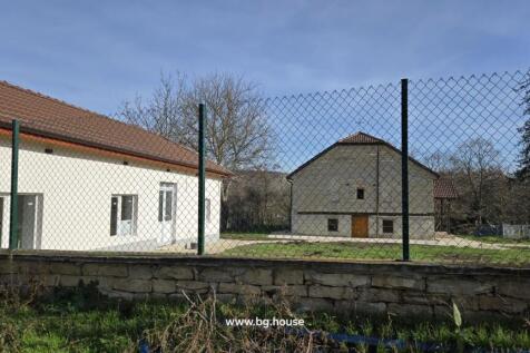 Exterior view showing two houses behind a wire fence with grass and trees in the foreground under a clear sky.