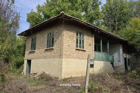 View of a traditional Bulgarian house showing stone lower walls and wooden upper walls with green window frames, surrounded by trees and garden vegetation.