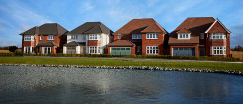 Modern suburban houses overlooking a lake with a paved pathway and well-kept gardens under a blue sk