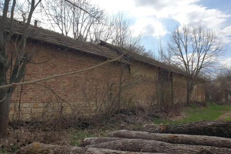 A long brick wall with a tiled roof runs alongside a natural garden area with stacked logs in the foreground and a cloudy sky above.