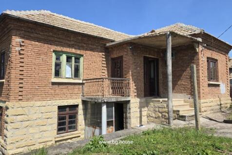 Front view of a traditional Bulgarian house showing stone base, brick walls, tiled roof, small balcony, and entrance porch