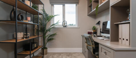 A cosy home office with a window, desk, computer, and decorative shelves filled with ornaments and p