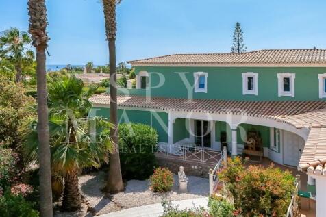 Exterior view of a green Algarvian luxury villa in Carvoeiro, showcasing traditional architecture, a paved garden path, palm trees, and spacious covered terraces.