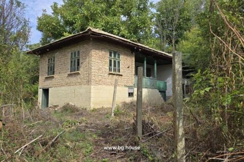 Side perspective of a traditional Bulgarian house featuring a stone cellar, wooden balcony with green railings, and garden area with trees and shrubs.