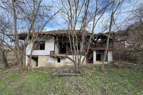 Exterior view of an old house in Balkanski village showing white walls, wooden balcony, and surrounding bare trees.