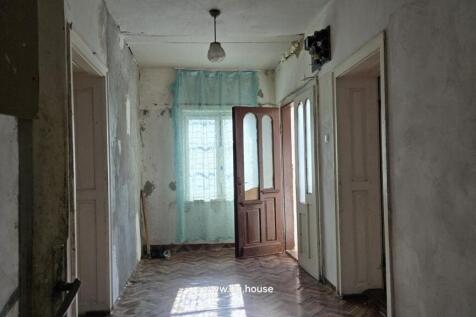 A corridor featuring aged walls, a wooden parquet floor, and two doors opening to adjacent rooms, illuminated by natural light through a curtained window.