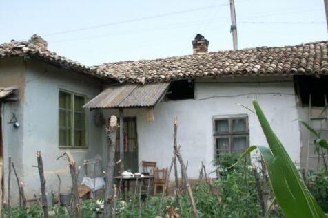 Exterior view of a traditional Bulgarian village house with a tiled roof and garden plants enclosed by a rustic wooden stick fence.