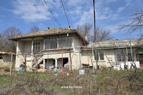 View of a two-storey Bulgarian house with a tiled roof, external stairs, and a garden area with dry grass and leafless trees