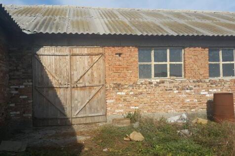 Exterior of a brick farm building showing wooden double doors, multiple windows, and a rusty barrel on the ground surrounded by dry grass and debris.