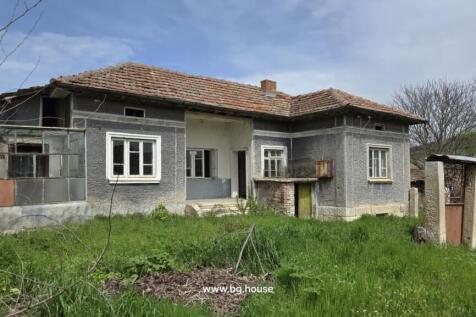 Front view of the Bulgarian rural house showing grey walls, tiled roof, and surrounding garden area