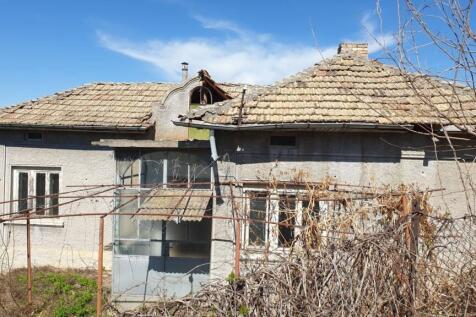 Side perspective of a grey village house featuring a tiled roof and an enclosed porch, with dry vegetation and bare branches around the property.