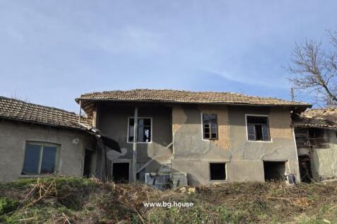 Exterior view of an old Bulgarian house with tiled roof and several windows, showing the rustic facade and surrounding dry vegetation.