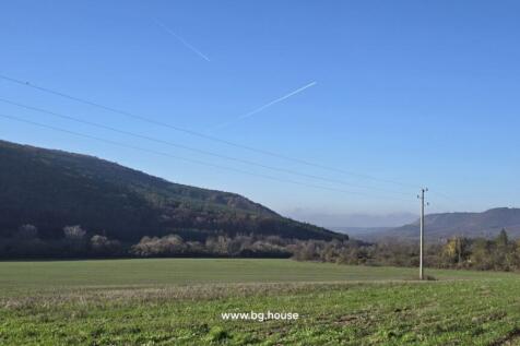 Wide open green field bordered by wooded hills under a clear blue sky with visible airplane contrails.