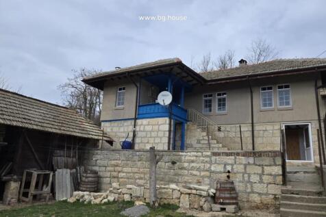 Side perspective of the Bulgarian house featuring a blue wooden balcony and stone stairs leading to the entrance