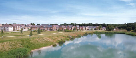 Modern houses beside a lake with a walking path, surrounded by trees and greenery under a blue sky a