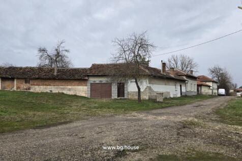 View of a corner Bulgarian house with tiled roof, stone base, and adjacent buildings at the end of a village street.