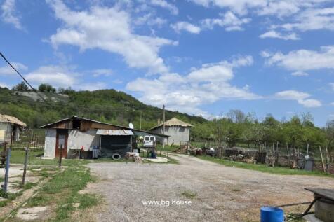 Farmyard scene showing small outbuildings, a dirt road, and green trees with hills in the background on a sunny day.