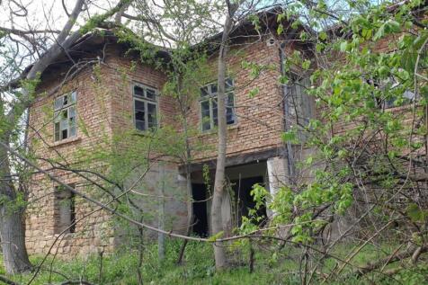 Two-storey rural house made of brick and stone with multiple windows, partially obscured by trees