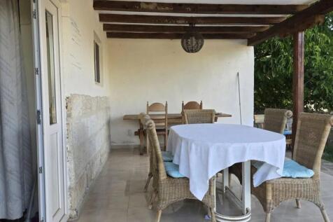 A covered terrace featuring a dining table with a white tablecloth surrounded by wicker chairs, adjacent to the house exterior wall.