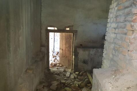 Interior room showing rough plaster walls, bricks scattered on the floor, and an open wooden door providing access to the outside.