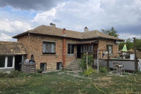 Exterior view of a brick house with a tiled roof, wooden porch, and garden area with grass and plants.