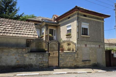 Front view of a traditional Bulgarian house with a tiled roof, stone wall, and metal gate.
