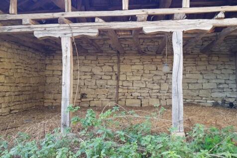 Exterior view of a stone outbuilding featuring wooden beams and an open side, with green plants growing nearby.