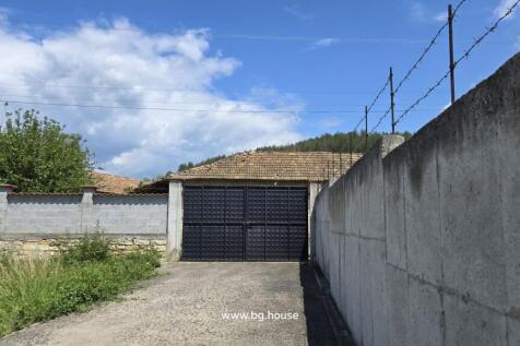 Entrance gate to the property with concrete boundary walls and a house roof visible behind under a blue sky with clouds.