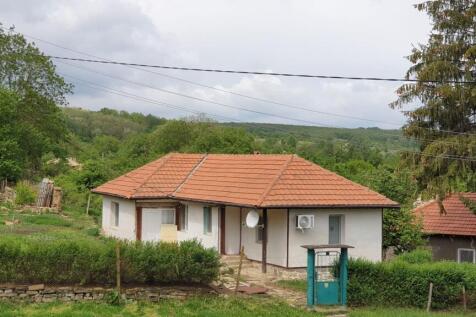 A white single-storey house with a terracotta tiled roof set in a green rural environment with trees and bushes.