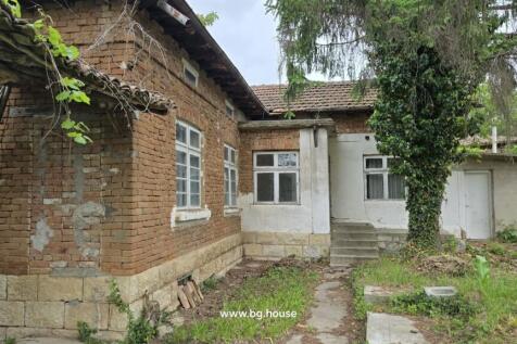 Exterior view of a traditional Bulgarian brick house with white window frames and a tiled roof, set in a garden with trees and plants.