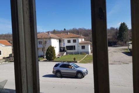 View looking out from a window showing a parked silver car on a village street with houses and trees.