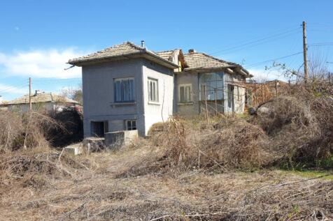 Exterior view of a traditional Bulgarian village house with a tiled roof and grey walls, surrounded by dry bushes and grass under a clear blue sky.
