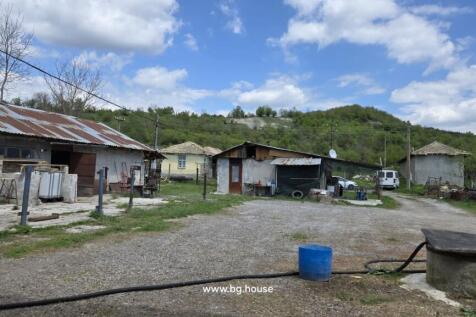 View of multiple farm buildings and a yard area with gravel and grass, surrounded by trees and hills.
