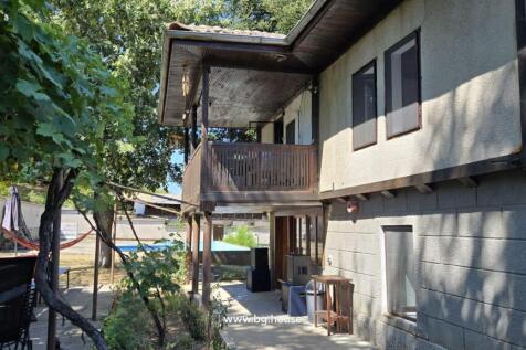 Side perspective of the house showing a wooden balcony above a shaded garden area furnished with tables and chairs.
