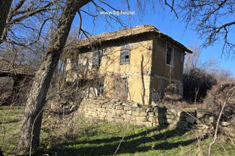 Exterior view of a two-storey rural house with wooden windows and a stone wall, surrounded by leafless trees under a clear blue sky.