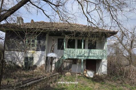 Exterior view of an old rural house featuring a tiled roof and a green wooden balcony with surrounding leafless trees and wire fence.