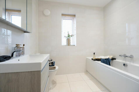 A modern bathroom with a white sink and toilet in the foreground and a white bathtub with a window b