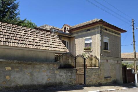 Side perspective of the Bulgarian house highlighting the tiled roof and stone wall with a metal gate.