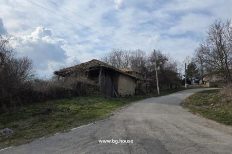 A paved rural road curves past old wooden and stone outbuildings with leafless trees and cloudy sky above.