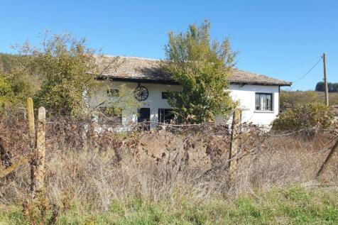 Exterior view of a white house with a tiled roof seen behind a wire fence and dry overgrown vegetation