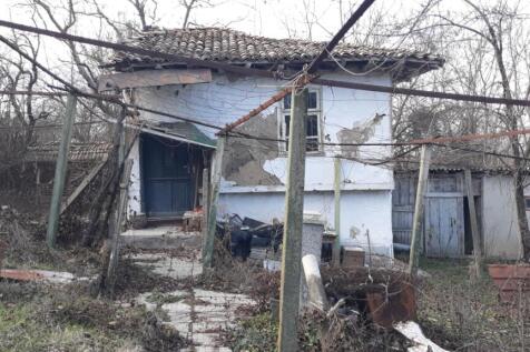 View of the front exterior of a traditional Bulgarian house showing an open doorway, tiled roof, and garden area with various items and bare trees