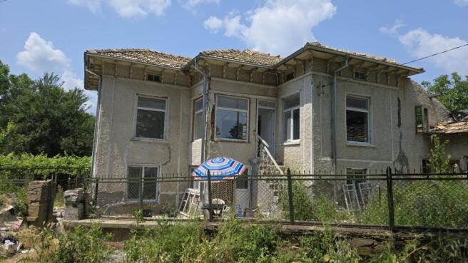 Front view of the two-storey Bulgarian house showing textured walls, multiple windows, and a small outdoor seating area with a striped umbrella