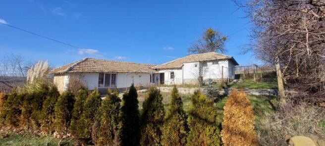 View of the white rural house with tiled roof and evergreen shrubs in front under a clear blue sky.
