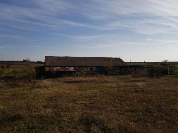 Wide view of the farm building with tiled roof and metal fencing, set in open grassy land under a partly cloudy sky.