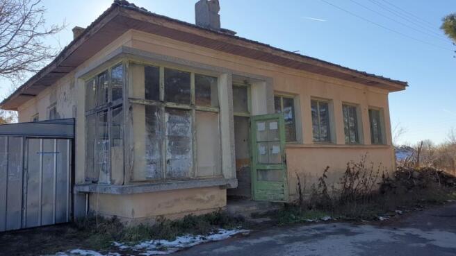 Side perspective of the old bakery showing beige walls, green door, and adjacent metal shed under a clear sky.